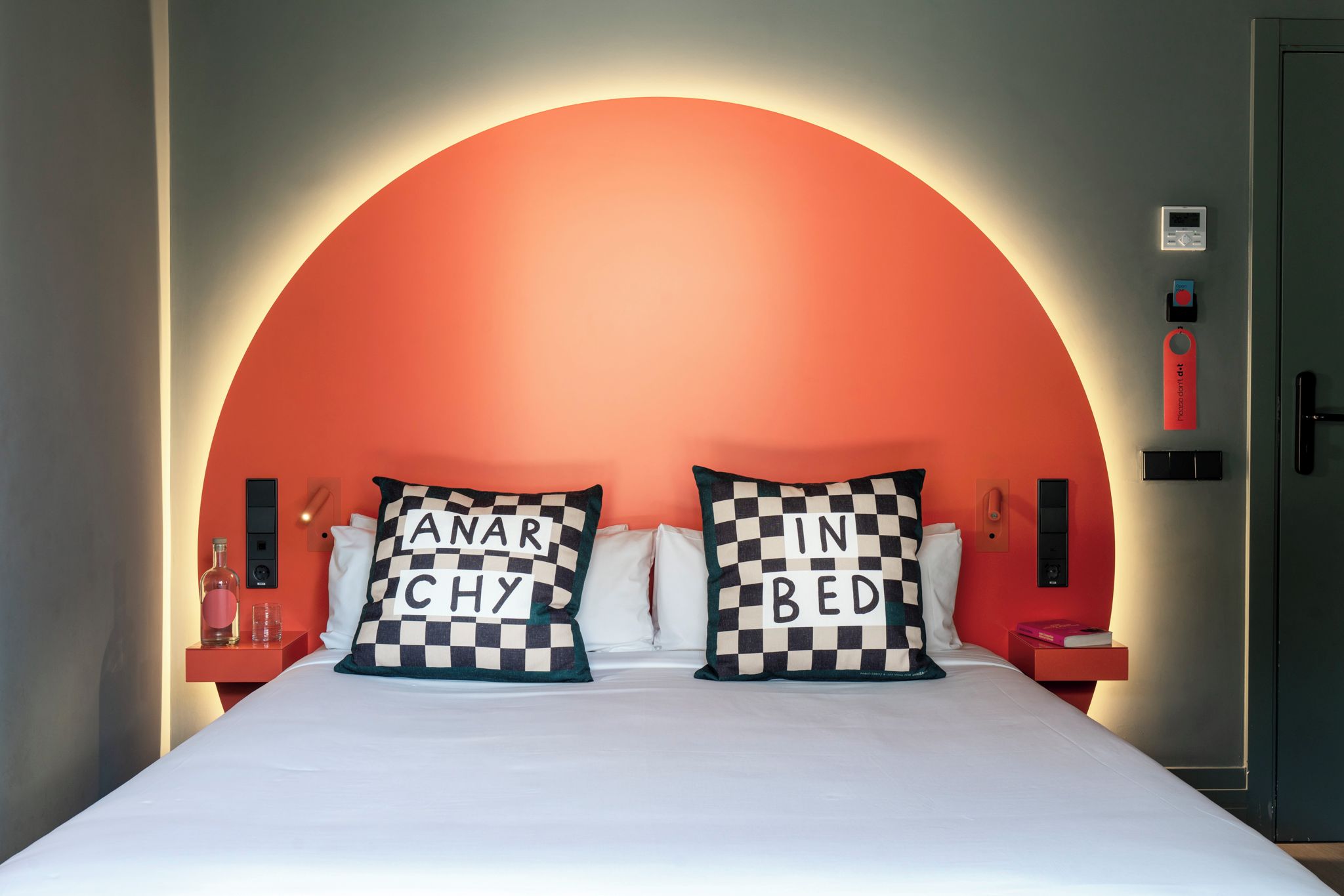 Una cama con dos almohadones a cuadros, una mesita de noche con una botella y vasos, y un panel de control en la pared.