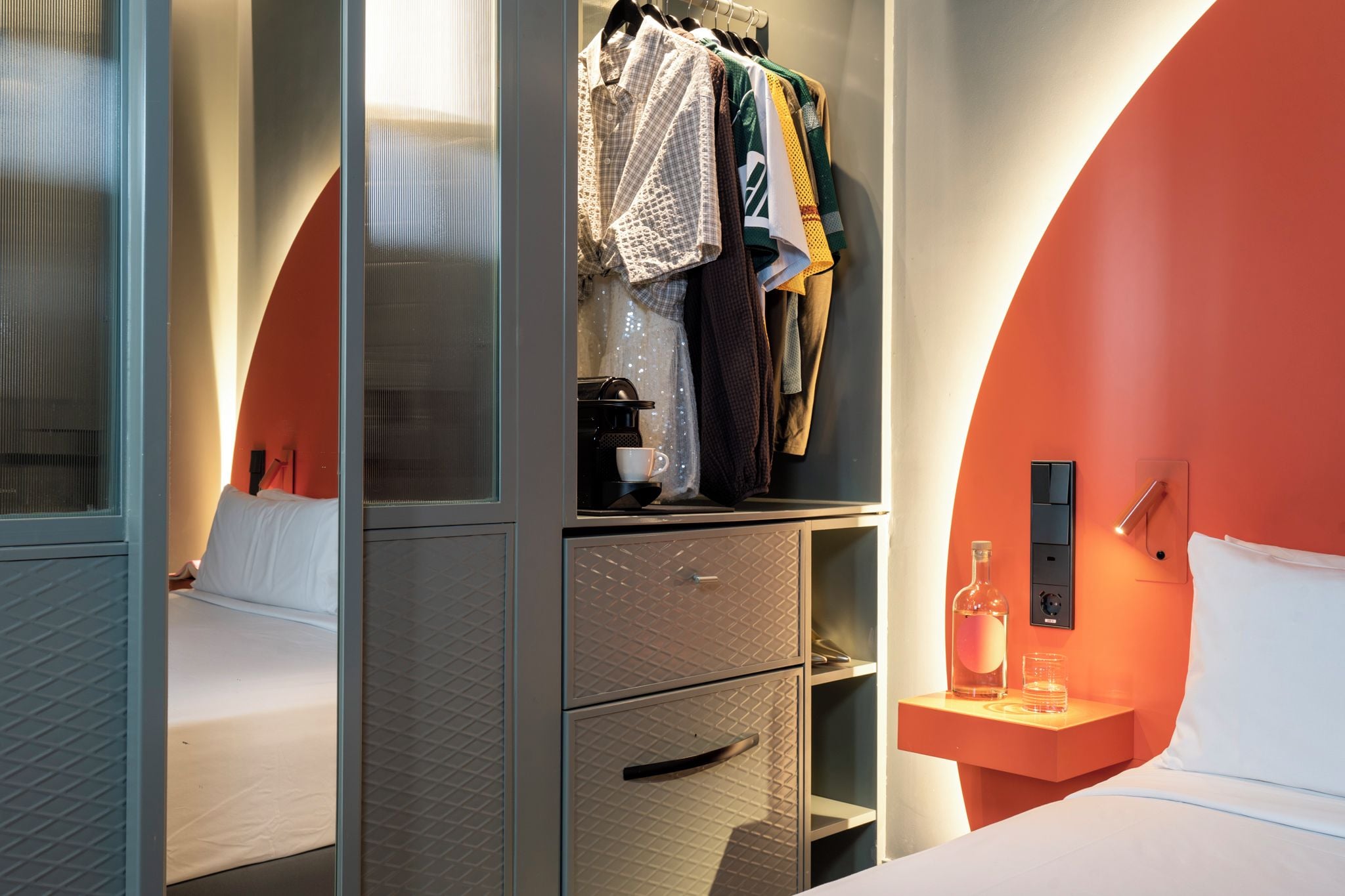 Quarto de hotel com cama, guarda-roupa com roupas penduradas, mesa de cabeceira com garrafa e copos, e luminária fixada na parede.
