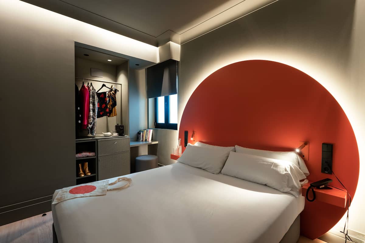 A modern bedroom with a large bed, a circular red accent wall, a bedside table with a phone, and a wardrobe with hanging clothes.
