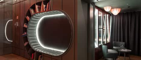 Una habitación de hotel moderna con una zona de estar curvada, una estantería, una pequeña mesa con un vaso y una ventana con cortinas.