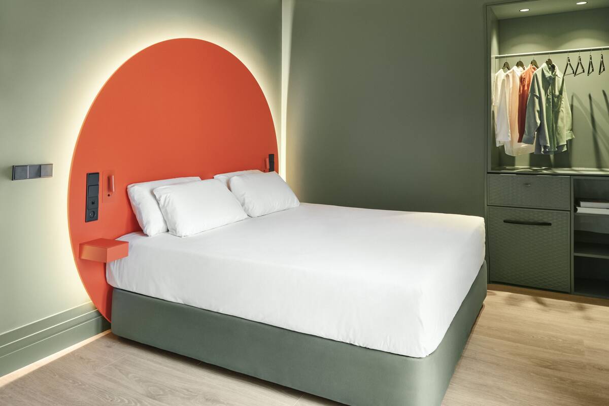 A modern bedroom with a curved orange headboard, a bed with white linens, a bedside table, and a built-in wardrobe with hanging clothes.