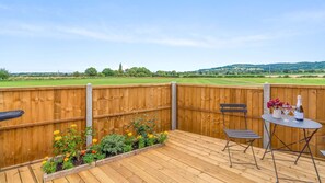A wooden deck with a table and chairs, a flower bed, and a view of a green field and hills in the distance. - Outbak Shepherds Hut - sleeps 2 guests in 1 bedroom (Hailes)