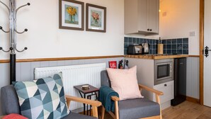 A cozy living room with a sofa, a coffee table, and a fireplace. There is a kitchen area with a stove, oven, and a tiled backsplash. - Outbak Shepherds Hut - sleeps 2 guests  in 1 bedroom (Hailes)