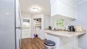 A compact kitchen with white cabinets, a wooden floor, a trash bin, and a window with blinds.