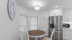 A modern kitchen with white cabinetry, a stainless steel refrigerator, a round wooden table with chairs, and a wall clock. - Summer House 202 by Deserving Vacations (Isle of Palms)