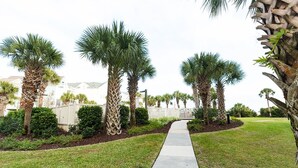 A pathway lined with palm trees and a white fence, leading to a building with a white facade and a green roof. - Summer House 202 by Deserving Vacations (Isle of Palms)