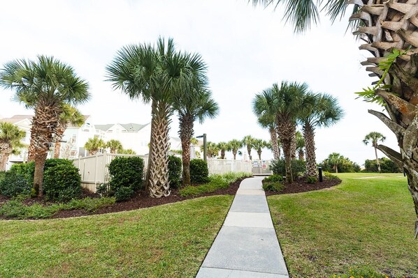 A pathway lined with palm trees and a white fence, leading to a building with a white facade and a green roof. - Summer House 202 by Deserving Vacations (Isle of Palms)