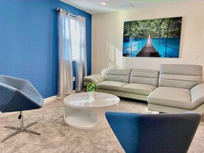 A living room with a blue wall, a beige sofa, a round coffee table, and a modern chair. - Luxury 7 Bdr~Private Pool at Champions Gate (Four Corners)
