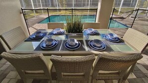 A glass-top dining table with wicker chairs, set for six, overlooking a swimming pool and neighboring houses.
