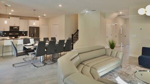 A modern living room with a large beige sectional sofa, a dining area with a black table and chairs, and a kitchen with white cabinets and stainless steel appliances.