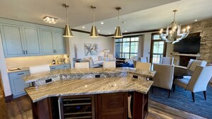 A spacious kitchen with granite countertops, a central island, and a stone fireplace. There are multiple pendant lights and a large TV mounted on the wall.