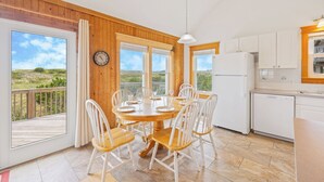 A dining area with a wooden table and white chairs, a kitchen with white cabinets, and a view of the outdoors through large windows. - Oceanfront Cottage with Grill, Deck, and Beach Only Steps Away. It's No Problem! (Avon)