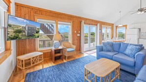 A spacious living room with a large TV, wicker furniture, a blue sofa, and a view of a house through the windows. - Oceanfront Cottage with Grill, Deck, and Beach Only Steps Away. It's No Problem! (Avon)
