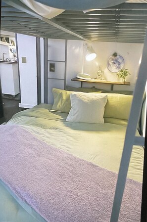 Quarto com cama, almofadas, mesa de cabeceira com abajur, prateleira com objetos decorativos e um prato fixado na parede.