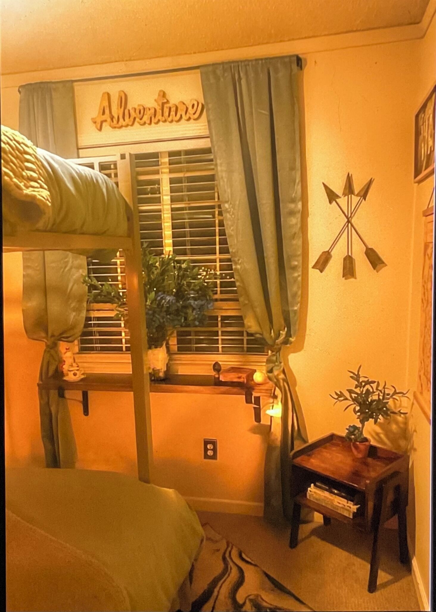 Une pièce chaleureuse avec un lit superposé, une fenêtre avec des rideaux, une petite table avec une plante et une décoration murale en forme de flèche.