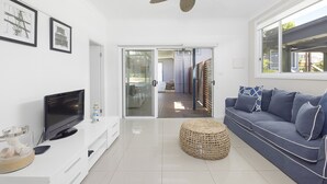 Uma sala de estar moderna com uma televisão de tela plana, um banquinho trançado, um sofá azul com almofadas estampadas e um ventilador de teto.