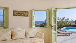 A cozy room with a floral sofa, a view of the sea through open windows, and a pool area.