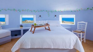 A bedroom with a large bed, a wooden bedside table with a tray, two windows with ocean views, a white chair, and a decorative vine border.