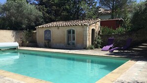Une piscine avec une terrasse carrelée, un petit bâtiment doté d’un toit en tuiles et deux chaises longues violettes.