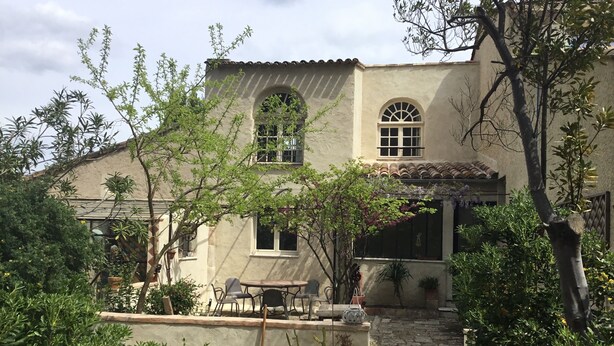 Un bâtiment doté d’un toit en tuiles, de fenêtres en arc de cercle et d’un chemin de pierre menant à un patio aménagé avec du mobilier d’extérieur.
