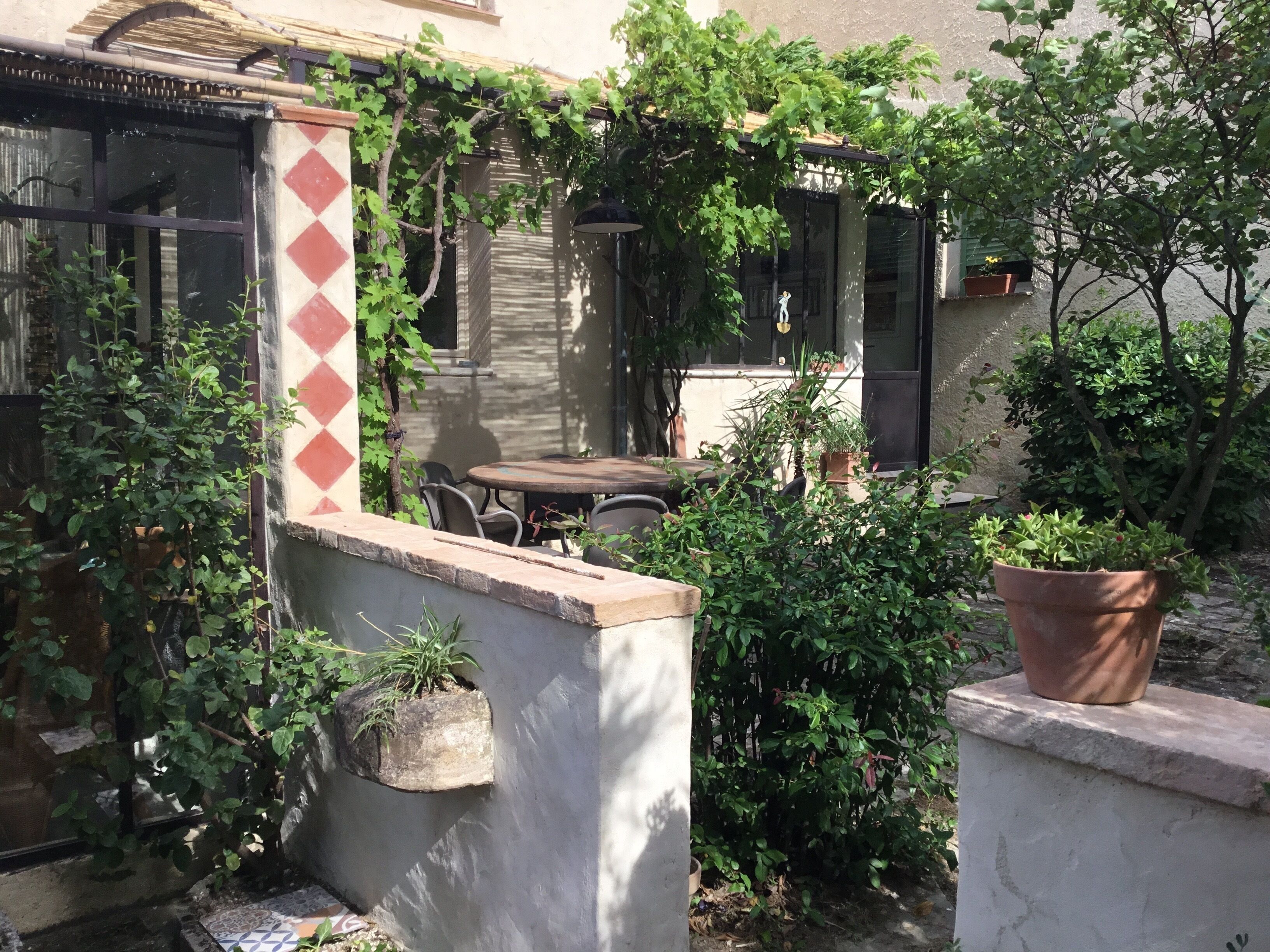 Un espace patio aménagé avec une table et des chaises, entouré de plantes en pot et d’un mur orné d’un motif géométrique.
