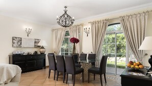 A dining area with a table and chairs, a chandelier, and a view of greenery through large windows. - Semi-detached house, 500m from the lake in Quinta do Lago, perfect for your vacation (Almancil)
