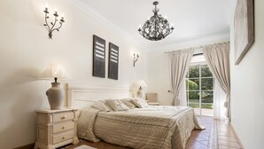 A bedroom with a bed, two bedside tables, a chandelier, a window with curtains, and a wall-mounted light fixture. - Semi-detached house, 500m from the lake in Quinta do Lago, perfect for your vacation (Almancil)