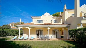 A large, two-story building with a tiled roof, a covered patio area with outdoor seating, and a well-maintained lawn. - Semi-detached house, 500m from the lake in Quinta do Lago, perfect for your vacation (Almancil)
