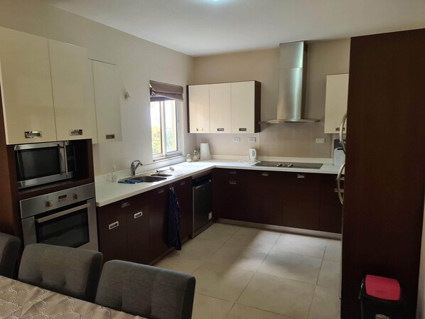 A compact kitchen with built-in appliances, a window, and a dining area with a chair. - Hausmann Follies Apartment (Kralendijk)