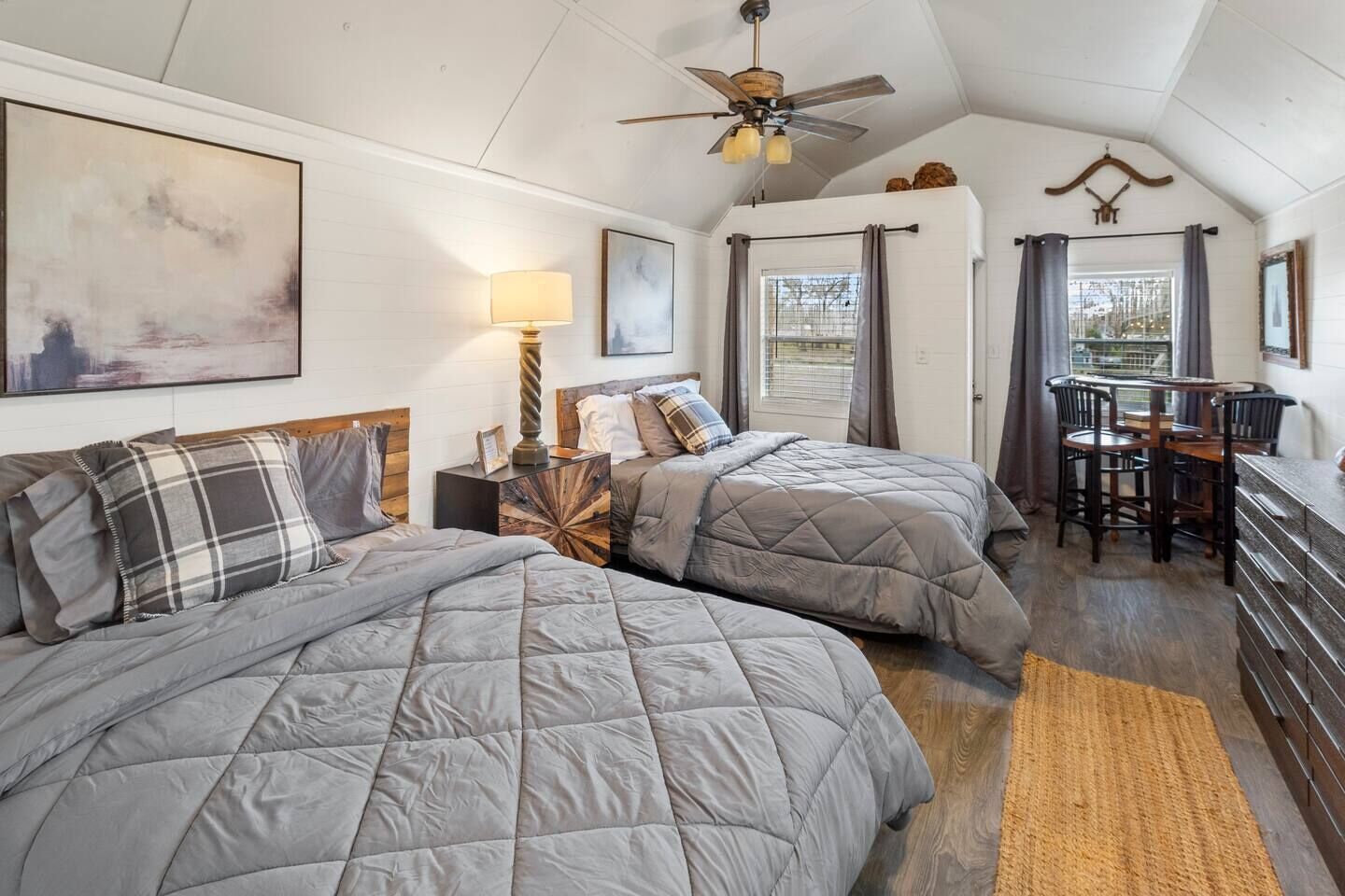 A bedroom with two beds, a ceiling fan, a desk with chairs, and a window with curtains.