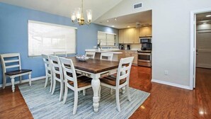 A dining area with a wooden table and white chairs, a chandelier, and a kitchen in the background. - Sandcastle Home (Sanibel)