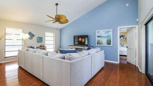 A living room with a sectional sofa, a ceiling fan, a television, and artwork on the walls. - Sandcastle Home (Sanibel)