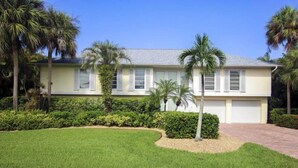 A two-story house with a light yellow exterior, white shutters, and a grey roof. There are palm trees and a well-maintained lawn in front. - Sandcastle Home (Sanibel)