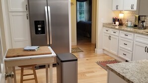 A compact kitchen with white cabinets, a stainless steel refrigerator, a small trash bin, and a wooden table.