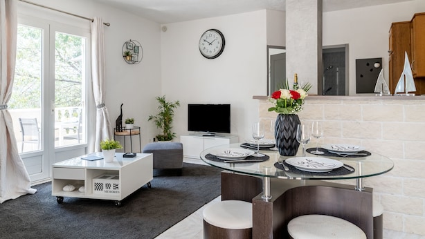 Ein modernes Wohnzimmer mit einem Glastisch für zwei Personen, einem Flachbildfernseher und einer gemütlichen Sitzecke mit einem weißen Sofa und Hocker.