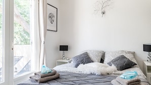 Ein ordentlich bezogenes Bett mit einer grauen Steudecke, zwei Kissen und einem weißen, flauschigen Kissen. Auf dem Bett liegen gefaltete Handtücher und an der Wand hängt ein gerahmtes Bild.