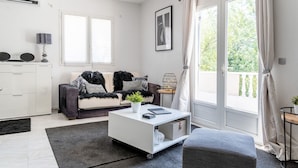 Ein modernes Wohnzimmer mit einem weißen Sofa, einem Couchtisch und einem Beistelltisch mit Lampe. Es gibt ein großes Fenster mit Vorhängen, eine Tür, die auf einen Balkon führt, und ein gerahmtes Bild an der Wand.