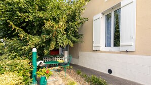 Un jardín con una mesa y sillas de metal verde, un regador verde y una pared blanca con una ventana.