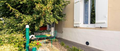 A garden area with a green metal table and chairs, a green watering can, and a white wall with a window.