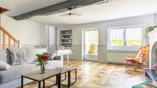 Una sala de estar con un sofá, una mesa de centro y una silla con un cojín colorido. Se observa una estantería y una ventana con cortinas.