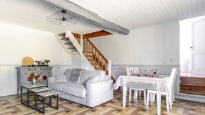 Amplia sala de estar con sofá, mesa de centro y comedor con sillas y una mesa dispuesta para una comida. Una escalera de madera conduce a un nivel superior.
