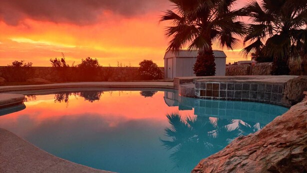A swimming pool with a clear view of the sunset, surrounded by rocks and palm trees. - Rancho Encantado VIP (El Paso)