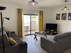 A living room with a sofa, a coffee table, a TV, and a balcony with yellow curtains.