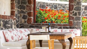 A stone-walled room with a wooden table, a cushioned bench, and a window overlooking a garden with red flowers.