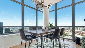 Ein Hotelzimmer in einem Hochhaus mit großem Tisch, vier Stühlen, einer Topfpflanze und weitreichender Stadtansicht durch bodentiefe Fenster.