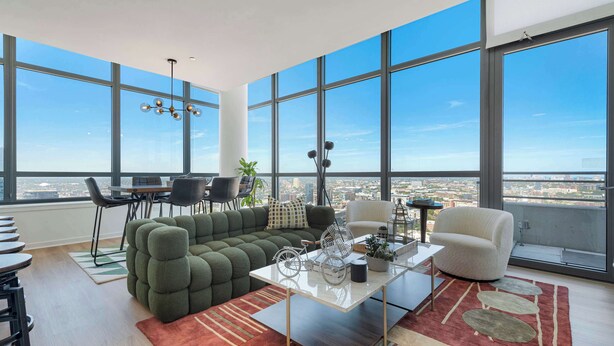 Ein modernes Wohnzimmer mit einem grünen Ecksofa, einem weißen Couchtisch und Blick auf die Stadt durch große Fenster.