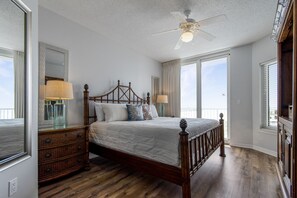 Une chambre avec un lit en bois, une commode, un ventilateur de plafond et de grandes fenêtres.