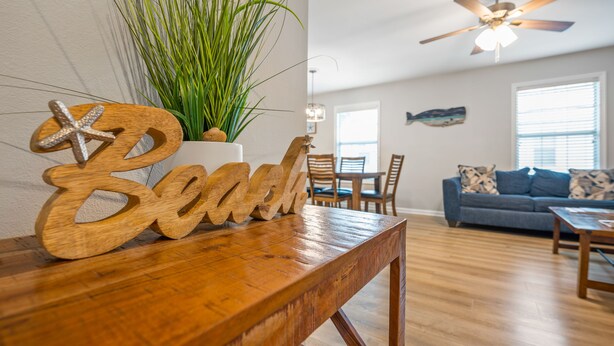 A wooden table with a starfish decoration and a wooden sign spelling "Beach." A potted plant with long green leaves is beside the table. A ceiling fan is visible in the room. - Beach Square – optional BIKES – Walk 2 Redfern Village, shops, and restaurants!. (St Simons Island)