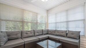 A sectional sofa with grey cushions and a glass-top coffee table in a sunroom with white blinds. - Beach Square – optional BIKES – Walk 2 Redfern Village, shops, and restaurants!. (St Simons Island)