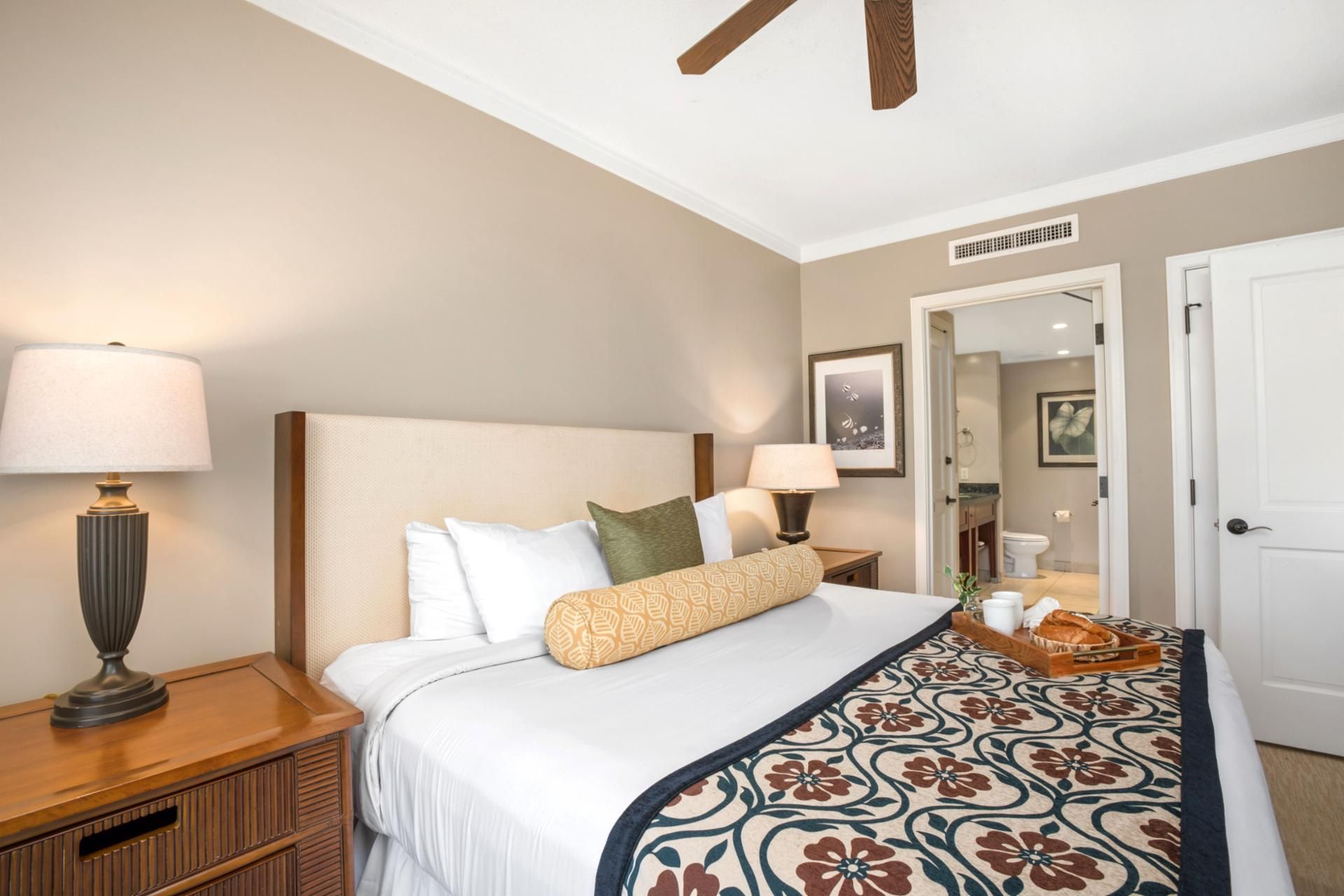 A neatly arranged hotel room with a large bed, two bedside lamps, a wooden nightstand, a tray with bread and a cup, and a door leading to a bathroom.